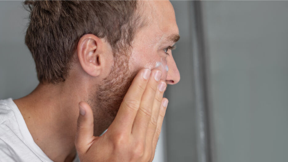 Ein Mann steht seitlich zur Kamera und cremt sich das Gesicht mit einer Anti-Aging-Pflege ein.